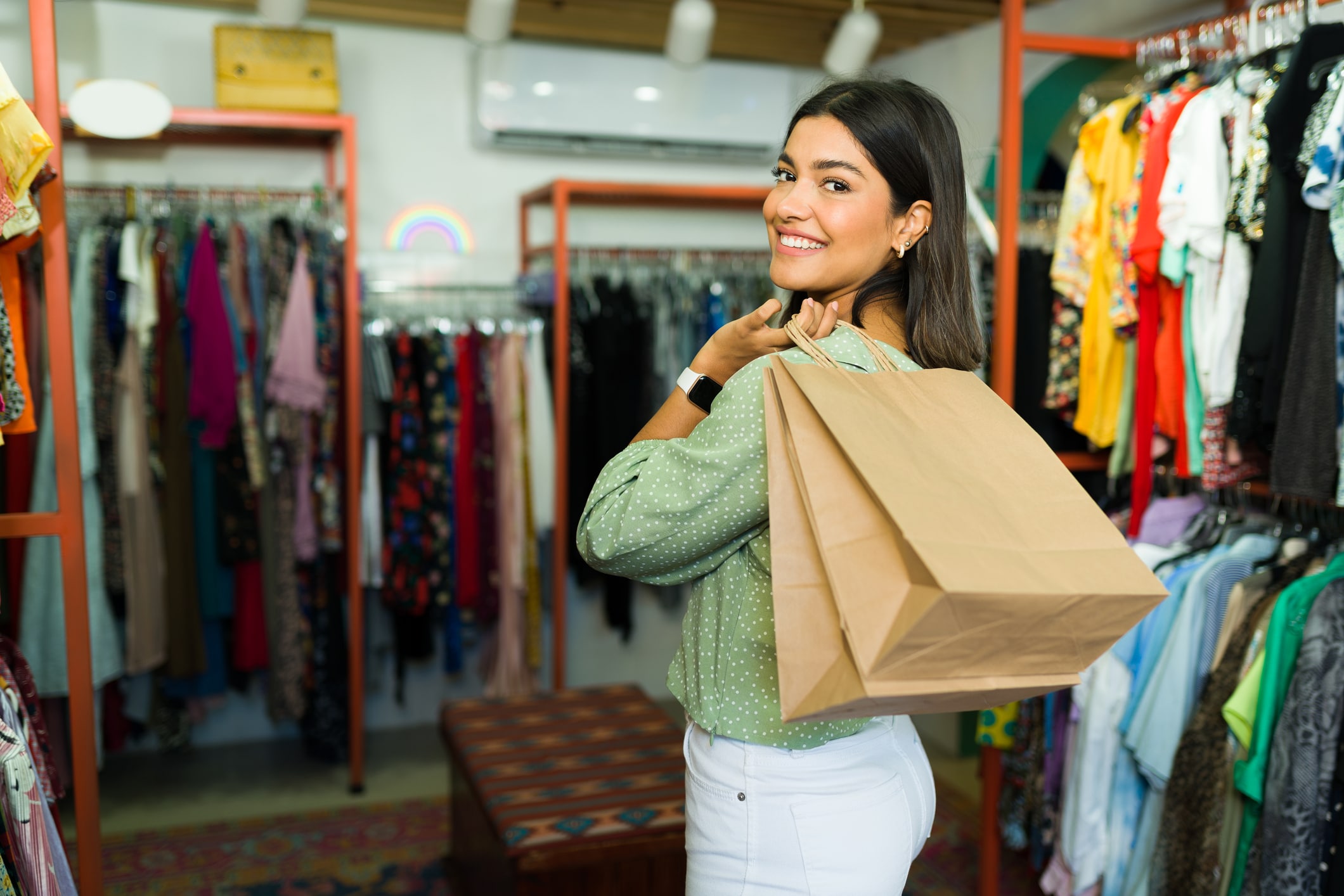 How to dress well on a budget. Woman with brown hair holding a shopping bag over her shoulder, with clothes on racks in the background.