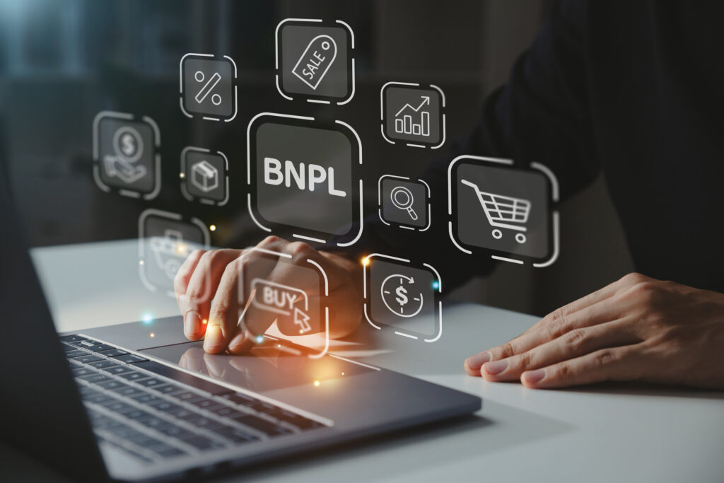 Buy now pay later companies, laptop computer with a hand on the mouse with icons showing BNPL, shopping cart, dollar sign, tag, percentage, and buy button.
