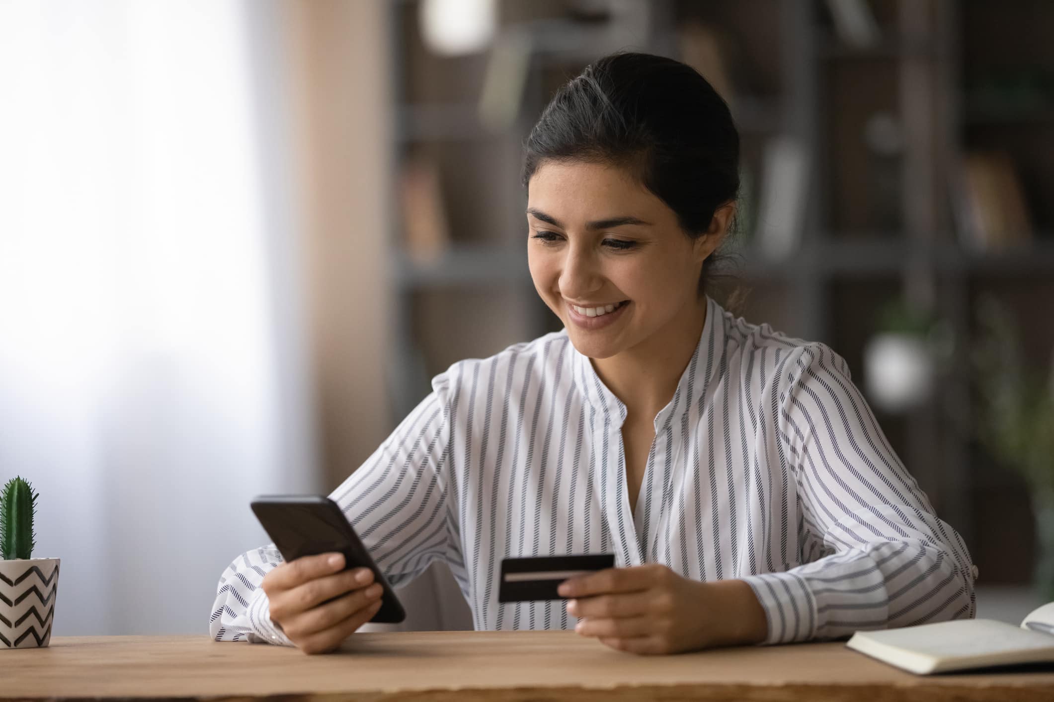 what credit score do you start with? Confident ethnic female student pay tuition fee remotely using mobile phone bank app. Smiling young indian lady sit at office table use credit card to make online money transfer buy goods at internet