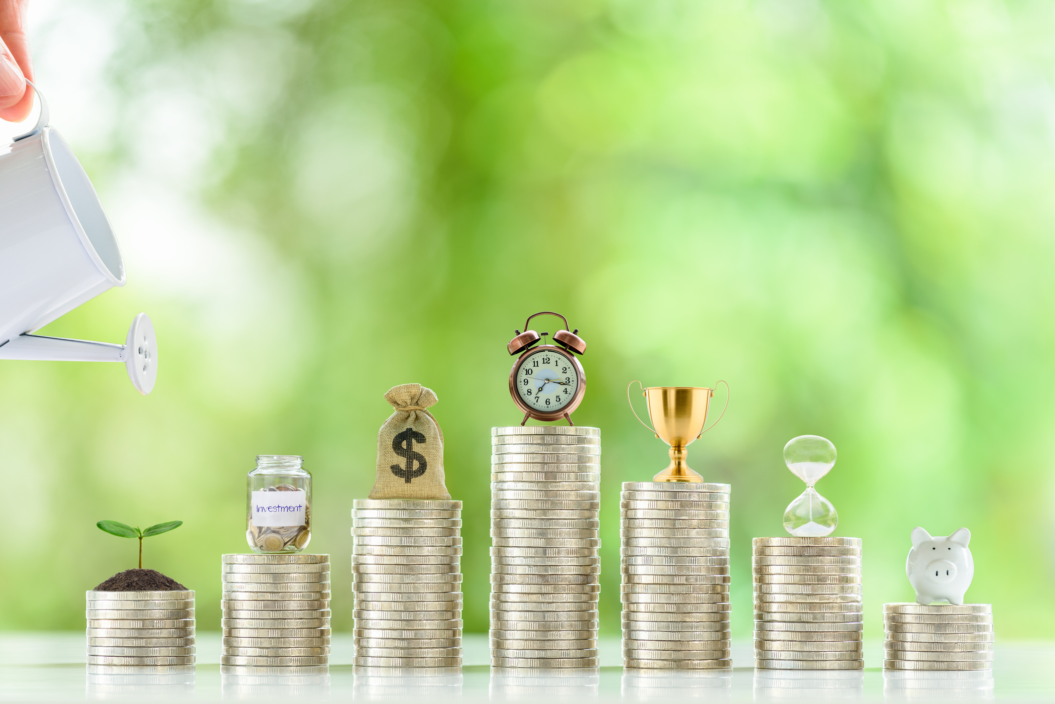 Long-term financial goal setting for most people, financial concept : Hand pours water from a watering can onto a young sprout or a small tree, an investment jar, a clock, US dollar bag on coin stacks